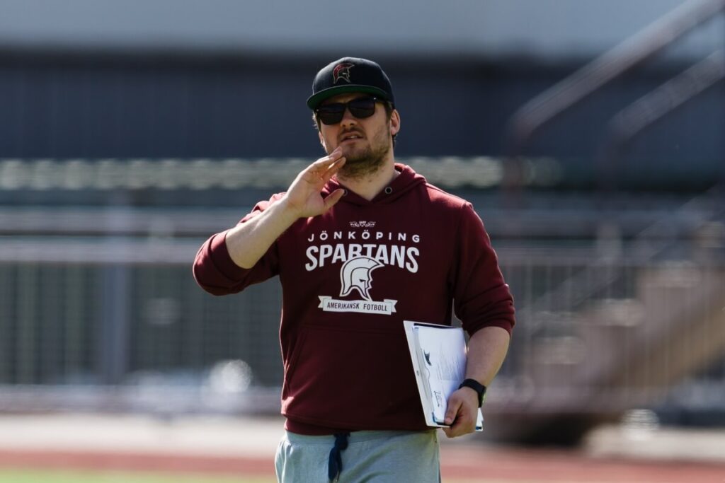 Coach wearing burgundy Spartans hoodie holds field clipboard.