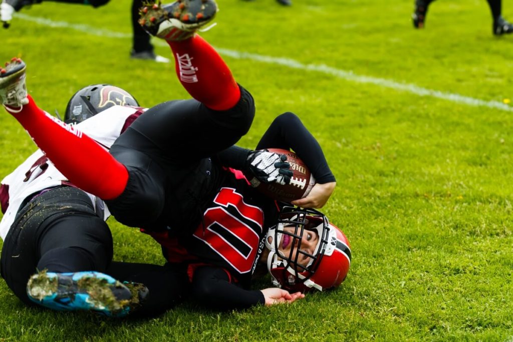 Red helmet football player tackles opponent on grass.