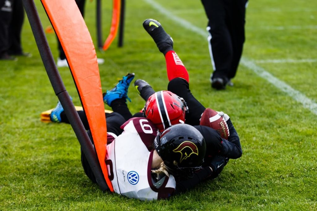 Football players tackle near orange field goal padding.