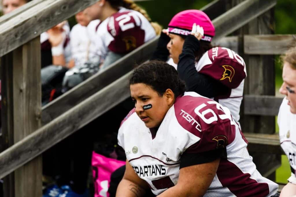 Female football players sitting on wooden bleachers.